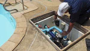 Swimming pool technician inspecting and repairing pipes, valves, and pump equipment inside an open pool maintenance compartment.